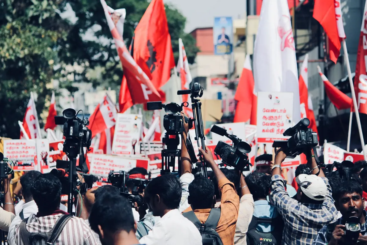 A vibrant protest in Chennai, India with media coverage featuring bright red flags and banners.