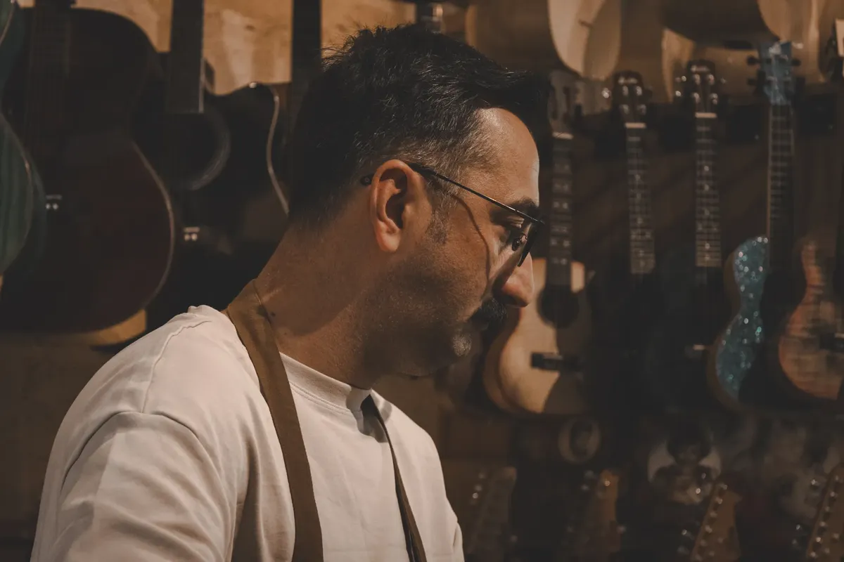 一名製琴師專注地工作，周圍擺滿了吉他和 Ukulele 在工藝工坊中。