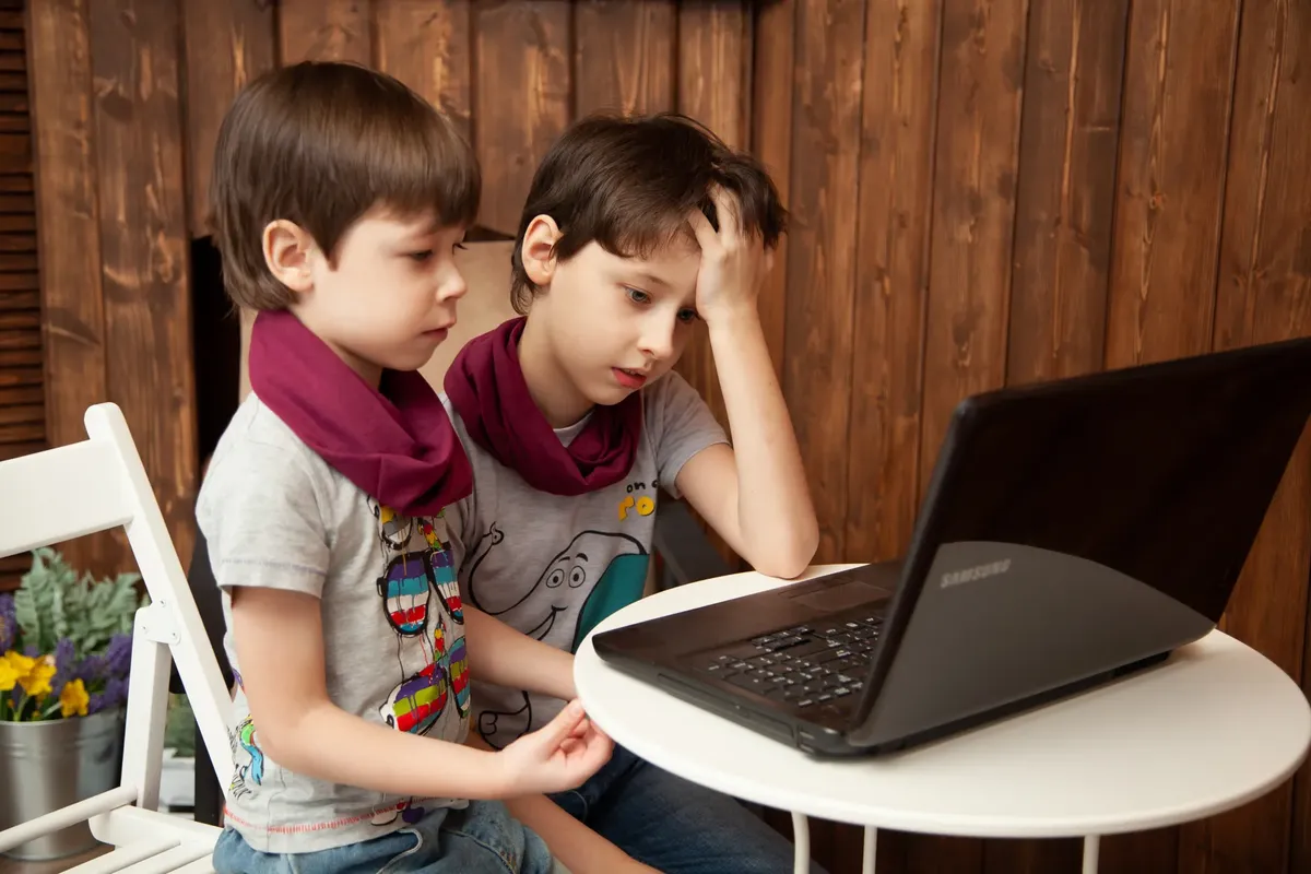 Two young boys intently watching content on a laptop indoors, wearing matching scarves.